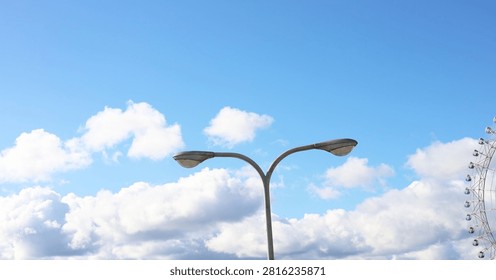 Low angle view of a double-headed street lamp post against a bright blue sky with fluffy white clouds and part of a Ferris wheel visible on the right