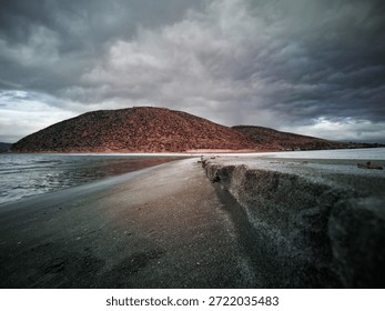 Low angle view of a dramatic lakeshore with textured sand, calm water and stormy clouds. Natural landscape capturing depth, atmosphere and quiet tension in nature. Salda Lake, Burdur.