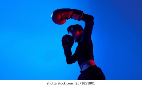 Low angle view dynamic image of young sportive woman delivering powerful punch with boxing gloves during training against blue studio background in neon light. Concept of sport, youth, street style