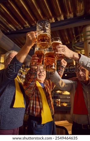 Low angle view of elderly men clinking mugs with lager beer. Friends gathering in modern pub, drinking beer and watching game translation. Concept of sport fans, elderly lifestyle, leisure activity