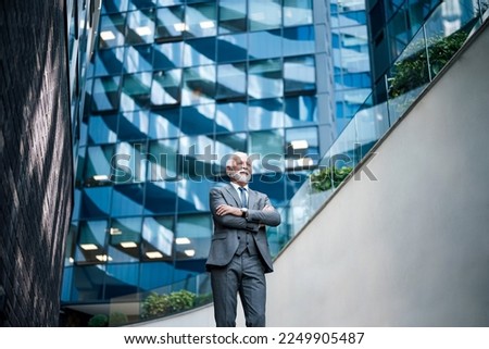 Low angle view of elderly professional looking away with arms crossed. Thoughtful senior businessman is standing outside modern business office building