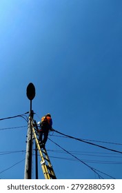 Low angle view of an electrician or lineworker wearing safety vest and helmet fixing power lines on a pole against a clear blue sky.