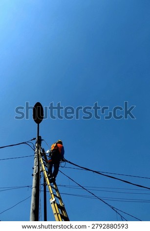 Low angle view of an electrician or lineworker wearing safety vest and helmet fixing power lines on a pole against a clear blue sky.