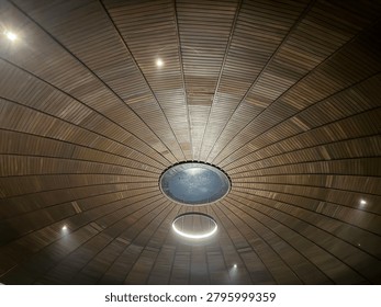 Low angle view of an elegant circular wooden dome ceiling with warm lighting. Contemporary architectural design featuring radial timber planks and a modern ring chandelier