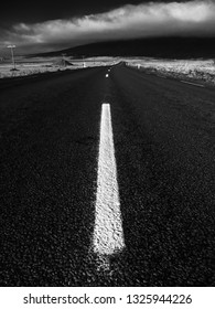 Low Angle View Of Empty Road Winding Into Distance With White Lines And Moody Clouds Over Mountains On Horizon.