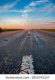 Low angle view of an empty rural asphalt road leading towards the horizon during sunset beneath a colourful dramatic sky, symbolising travel, freedom, direction, opportunity and new beginnings.