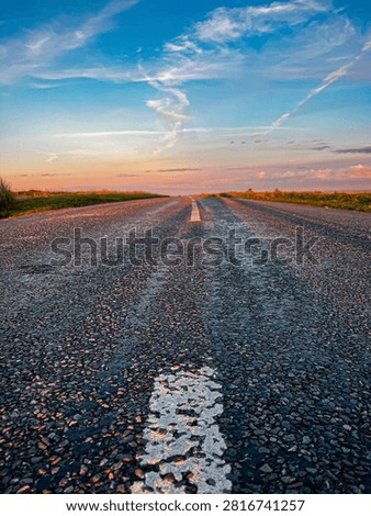 Low angle view of an empty rural asphalt road leading towards the horizon during sunset beneath a colourful dramatic sky, symbolising travel, freedom, direction, opportunity and new beginnings.
