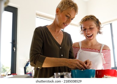 Low Angle View Of Excited Teenage Daughter With Mouth Open Laughing And Looking At Mother Preparing Food In Kitchen At Home