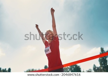 Low angle view of excited young female athlete with arms raised reaching the finish line at track field during marathon outdoors. Sports, motivation concept