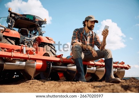 Low angle view of farm worker texting on cell phone while relaxing during soil cultivation in the field.