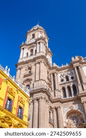 Low angle view featuring the historic Malaga Cathedral bell tower and tower clock next to a vibrant yellow and red residential building under a bright blue sky in Andalusia region, Spain.