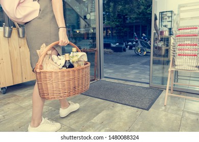 Low Angle View Of Female Walking Through A Grocery Store Door And Carrying A Basket Full Of Products.