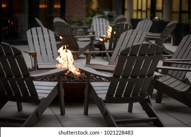 Low angle view of a fire pit surrounded by a circle of Adirondack chairs, with another campfire and hotel building in the dark background