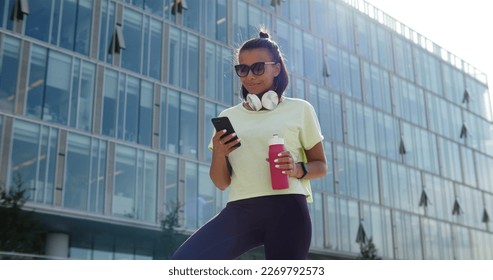 Low angle view fitness woman using phone with workout app exercising in city. Portrait of Indian female with bottle of water and smartphone after workout outdoors - Powered by Shutterstock