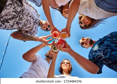 Low angle view of friends having fun at poolside summer party, clinking glasses with colorful summer cocktails near hotel swimming pool. People toast drinking fresh juice at luxury villa on vacation.