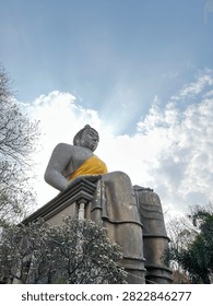 Low angle view of a giant sitting stone Buddha statue wearing yellow robe against a bright sky with sun rays.