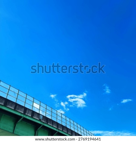 Low angle view of a green elevated highway sound barrier and lush trees against a bright blue sky with white clouds