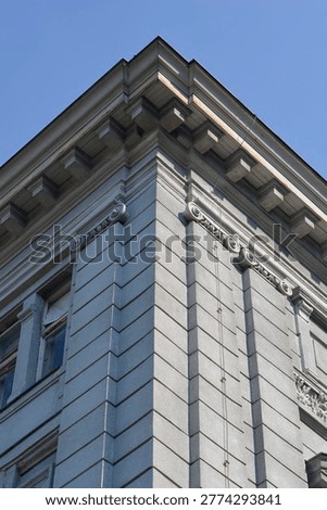A low angle view of a grey classical building corner featuring decorative cornices and ionic order details against a clear blue sky