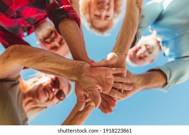 Low Angle View Of Group Of Cheerful Active Senior People Enjoying Sunny Summer Day Outdoors, Gathered In Circle, Holding Hands All Together In The Middle