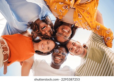 Low angle view of a group of five multiracial teenagers smiling and looking down at camera outdoors. Young student people of diverse ethnic cultures embracing while standing in a circle,together. High