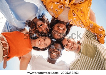 Low angle view of a group of five multiracial teenagers smiling and looking down at camera outdoors. Young student people of diverse ethnic cultures embracing while standing in a circle,together. High