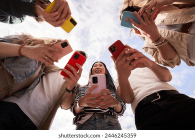 Low angle view of group happy unrecognizable people in circle using colorful mobiles phones together outdoor. Young friends and smiling Asian woman in center holding cells. Gen z technology addiction - Powered by Shutterstock