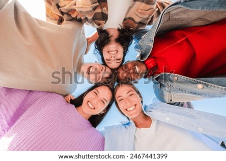 Low angle view of a group of multiracial young friends hugging in a circle, smiling and looking at camera with a positive expression. Team of teenagers laughing and ambrecing each others outside. High