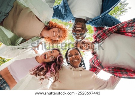 Low angle view of a group of multiracial happy young teenagers hugging each others in a circle, looking down at camera with smiling faces, representing friendship, diversity, and togetherness outdoors