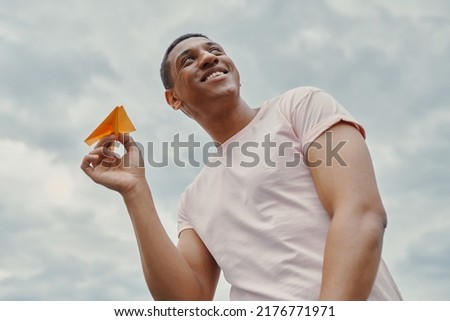 Low angle view of handsome african man holding paper airplane with sky in the background                  