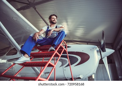 Low angle view of handsome bearded mechanic in uniform looking away and smiling while sitting near the aircraft in hangar