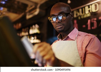 Low Angle View Of Handsome Black Male Waiter In Glasses Using POS System While Working In Restaurant