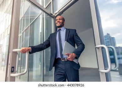 Low angle view of handsome young Afro American businessman in classic suit holding a laptop and smiling while leaving the office building - Powered by Shutterstock