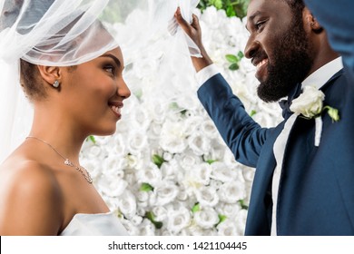 low angle view of happy african american bridegroom touching white veil and smiling near bride and flowers 