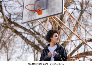 Low Angle View Of Happy African American Woman In Jacket Standing On Basketball Outdoor Court