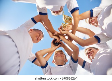 Low Angle View Of Happy  Boys In Junior Football Team Holding Trophy Cup Together Against Clear Blue Sky And Cheering