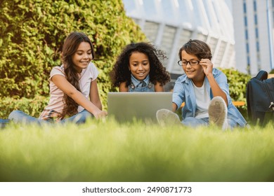 Low angle view of happy little preteen school children kids using digital wireless gadgets laptop with backpacks looking at screen sitting at the grass park outdoors doing home tasks project together - Powered by Shutterstock