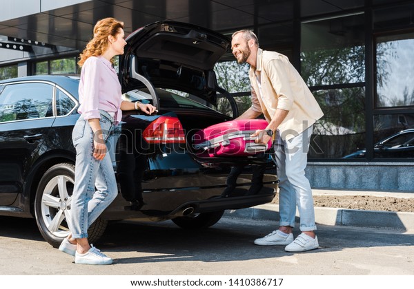 low angle view of happy man putting pink luggage\
in car trunk near woman 