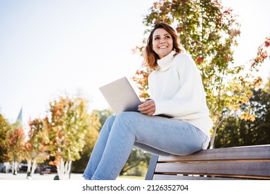 Low Angle View Of A Happy Vivacious Woman Using A Laptop Computer As She Perches On Top Of The Back Of A Wooden Bench In An Urban Park In Autumn