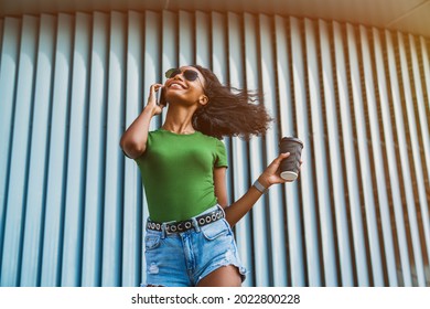 Low angle view of happy woman standing near wall with coffee while talking on mobile phone. Smiling african-american girl texting typing online messages, ordering food delivery outdoors. - Powered by Shutterstock