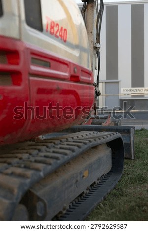 Low angle view of a heavy tracked excavator with a worn steel track in the foreground, parked beside a modern industrial building with grass and machinery