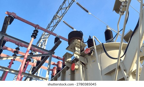 Low angle view of a high voltage power transformer and porcelain insulators at an electrical substation against a clear blue sky. Industrial energy distribution and electricity transmission infrastructure.
