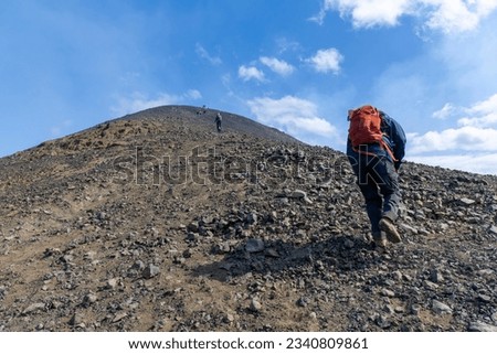 Low angle view of hikers on steep path to top of mountain Litli-Hrutur on Reykjanes Peninsula, Iceland, near fissure Litli-Hrútur Hill during the 2023 eruption against a white clouded blue sky