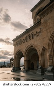Low angle view of the Holy Trinity Cathedral of Tbilisi (Sameba) at sunset. Majestic Georgian Orthodox architecture with golden stone facade under a dramatic evening sky in Georgia.