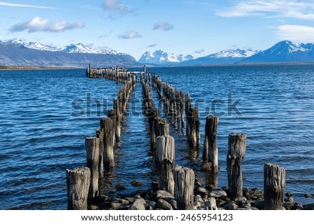 Low angle view of iconic poles of historical pier in the waters of Canal Senoret in front of Puerto Natales, Chile stretching into the water with snowcapped mountains in background