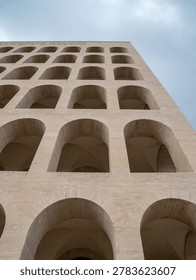 Low angle view of the imposing beige stone facade of the Palazzo della Civilta Italiana featuring symmetrical geometric arches under a cloudy sky in Rome showcasing historic rationalist architecture