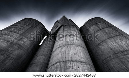 Low angle view of imposing concrete silos with a dramatic, dark, stormy sky overhead, evoking a sense of industrial power and foreboding.