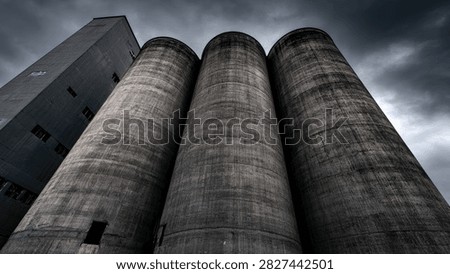 Low angle view of imposing concrete silos and a tall building under a dark, cloudy sky, evoking a sense of industrial power and foreboding.