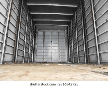 Low angle view inside empty cargo truck with metal walls, plywood floor, and scattered debris. Transportation, logistics, and shipping. Industrial background with perspective and symmetry 
Wide angle shot of empty box truck cargo area with corrugated metal walls and wooden floor. Some trash and dust visible. Transportation, logistics, shipping, and moving industry background.