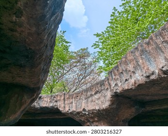 Low angle view from inside a rocky cave looking up at green trees and a clear blue sky. A perspective view looking up from the opening of an ancient stone cave towards the bright blue sky and lush green trees.