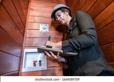 Low Angle View Of Inspector Woman Taking Notes During Energy Efficient Home Inspection, 220v Outlet And Water Valves On Wooden Wall In The Background